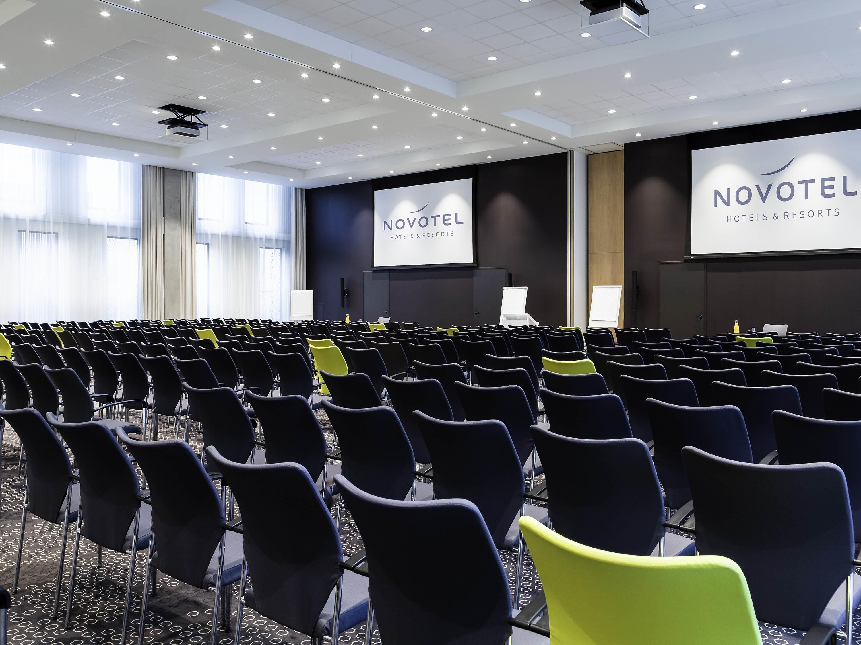 Une salle de conférence avec des rangées de chaises, un écran de projection et un écran de présentation affichant le texte « NOVOTEL HOTELS & RESORTS ».