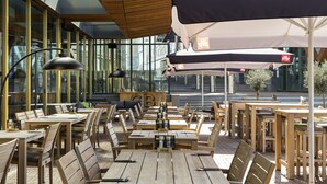 An outdoor cafe with wooden tables and chairs, umbrellas, and a view of the city through large windows.