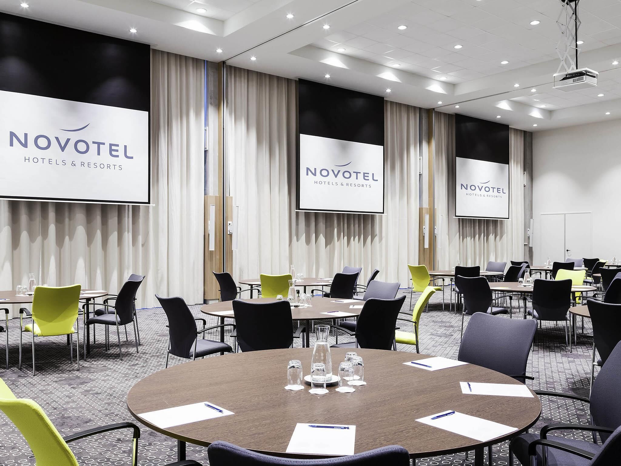 Un salón de conferencias con mesas, sillas y una pantalla de proyector que muestra 'NOVOTEL HOTELS & RESORTS'.