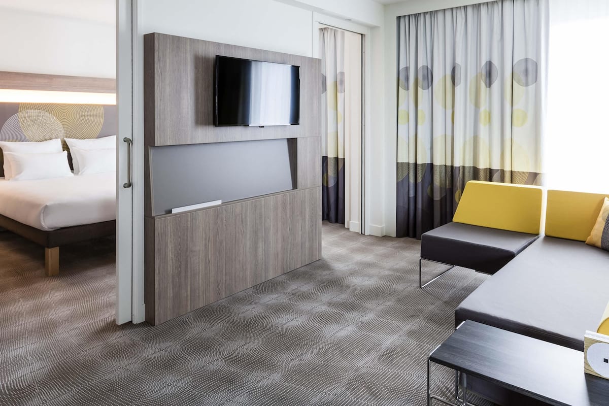 Una habitación de hotel con cama, televisor montado en la pared, escritorio con silla y ventana con cortinas.
