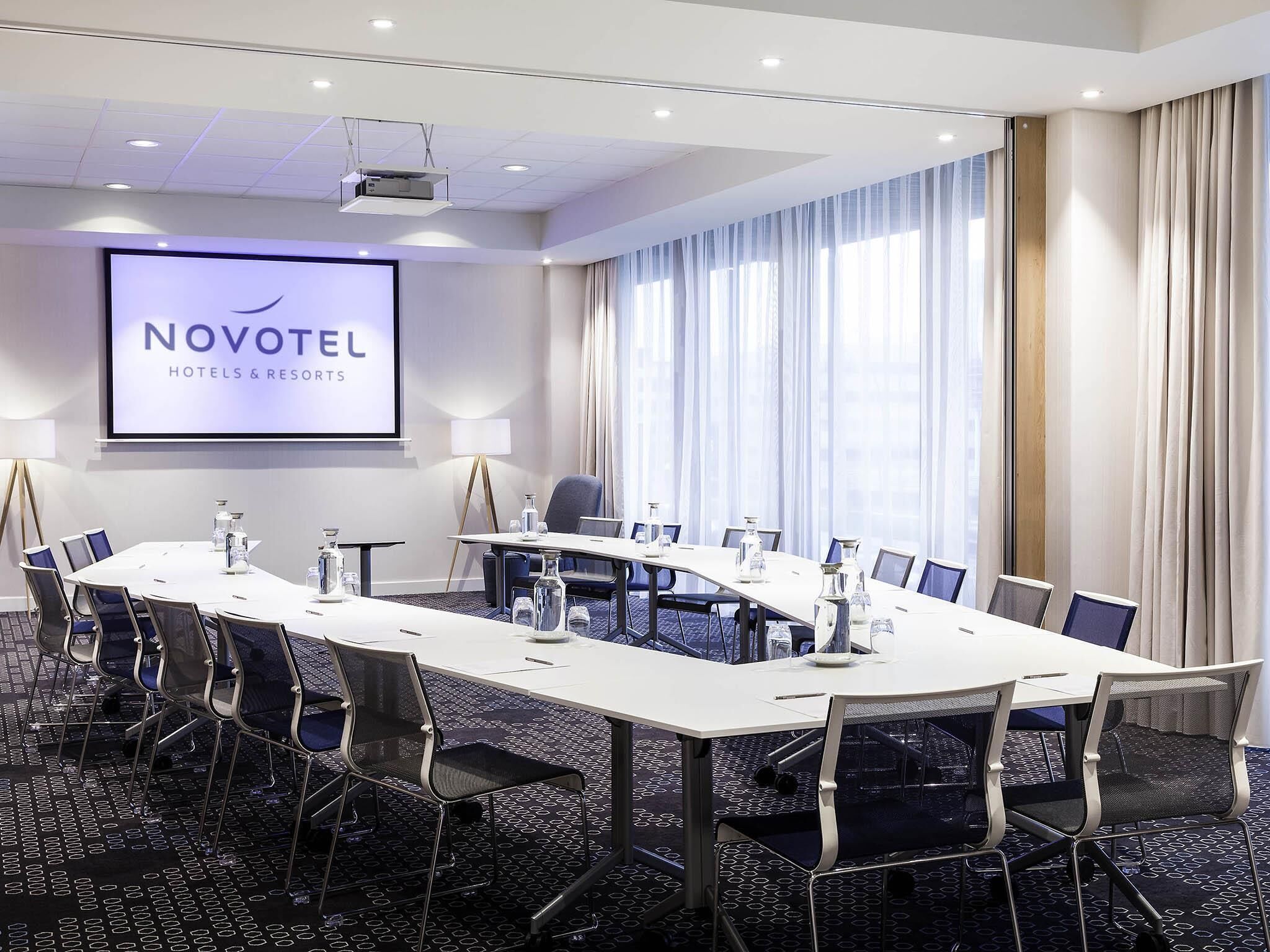 A conference room with a long table, chairs, a projector screen, and a large window with curtains.