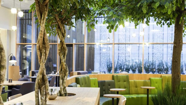 An indoor seating area with wooden tables, green cushioned chairs, and a large tree.