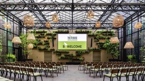 A spacious indoor venue with a high ceiling, featuring a stage with a 'WELCOME' sign, rows of chairs, and lush vertical gardens.