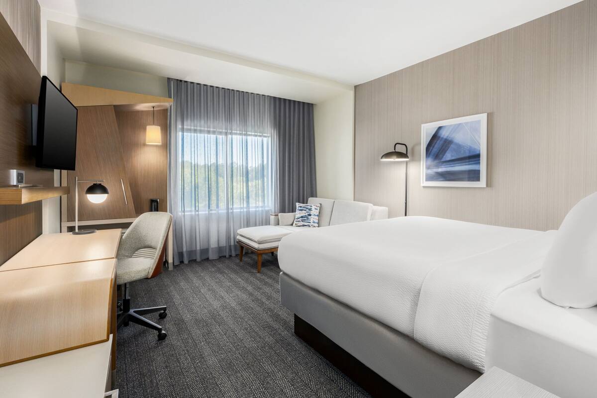 Ein Hotelzimmer mit einem großen Bett, einem Schreibtisch, einem Sessel, einem Fernseher, einer Lampe und einem Fenster mit Vorhängen.