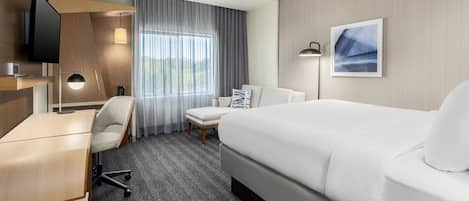 Habitación de hotel con una cama grande, un escritorio, una silla, un televisor, una lámpara y una ventana con cortinas.