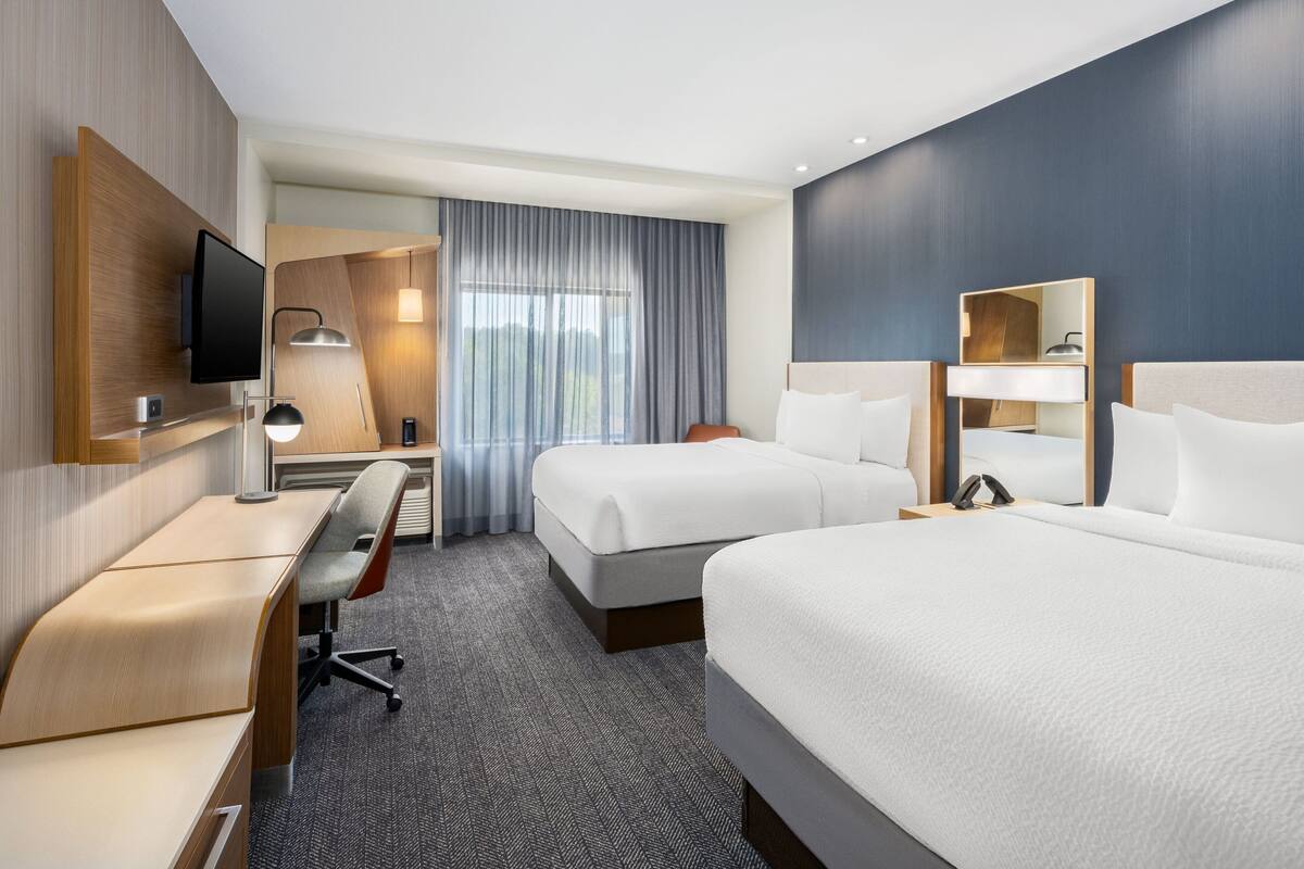 Ein Hotelzimmer mit zwei Betten, einem Schreibtisch, einem Stuhl, einem Fernseher und einem großen Fenster mit Vorhängen.