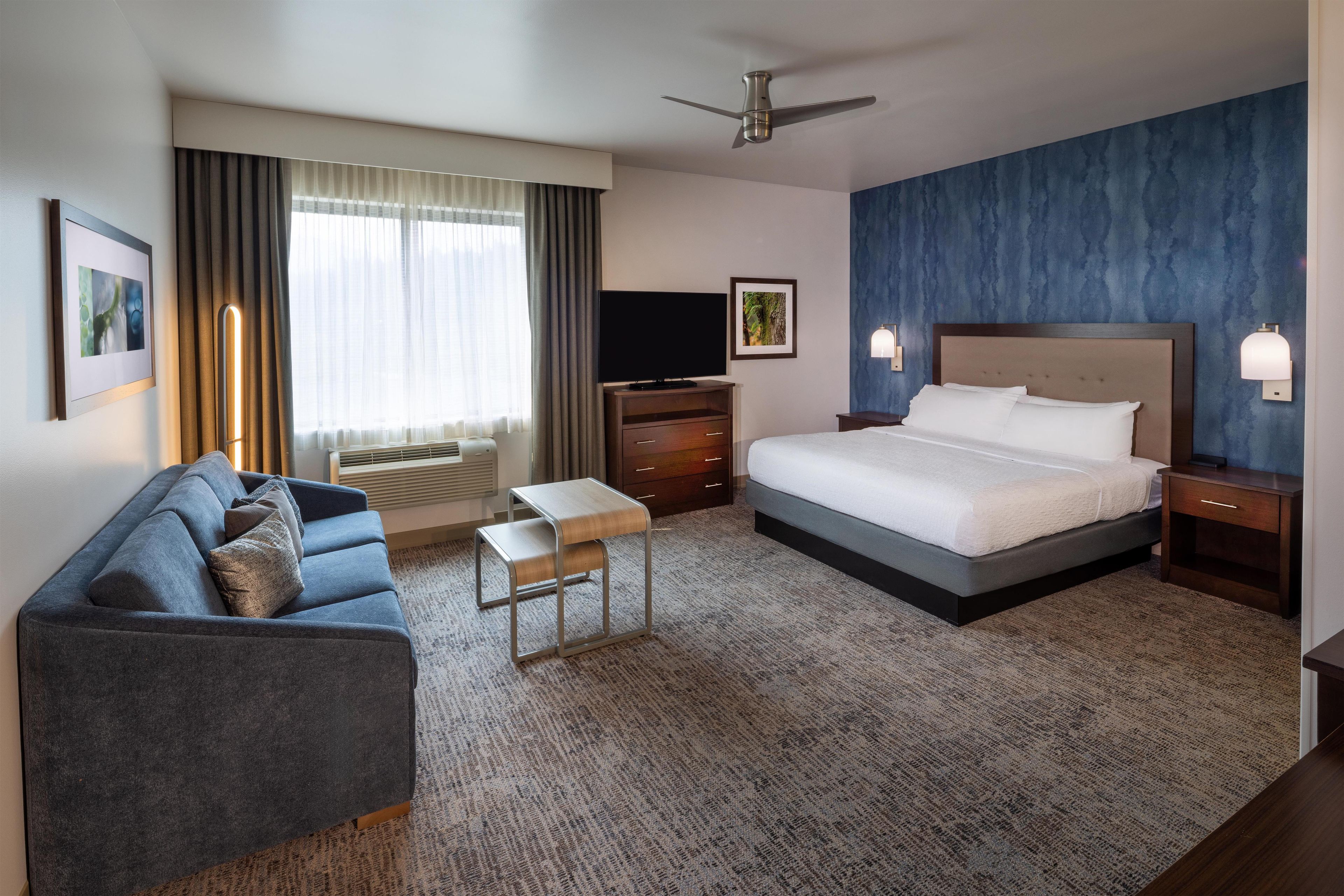 A hotel room with a bed, a sofa, a TV, a ceiling fan, and a large window with curtains.