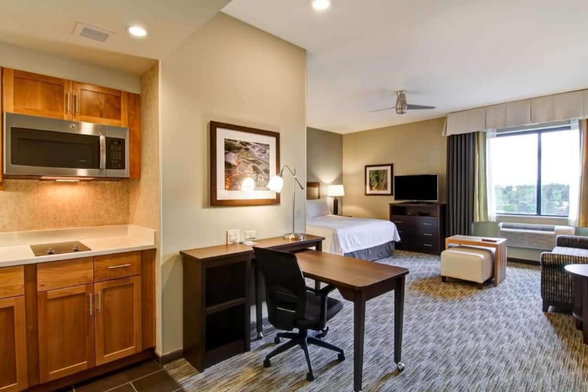 A hotel room with a kitchenette, a bed, a desk, a chair, a TV, and a window with curtains.