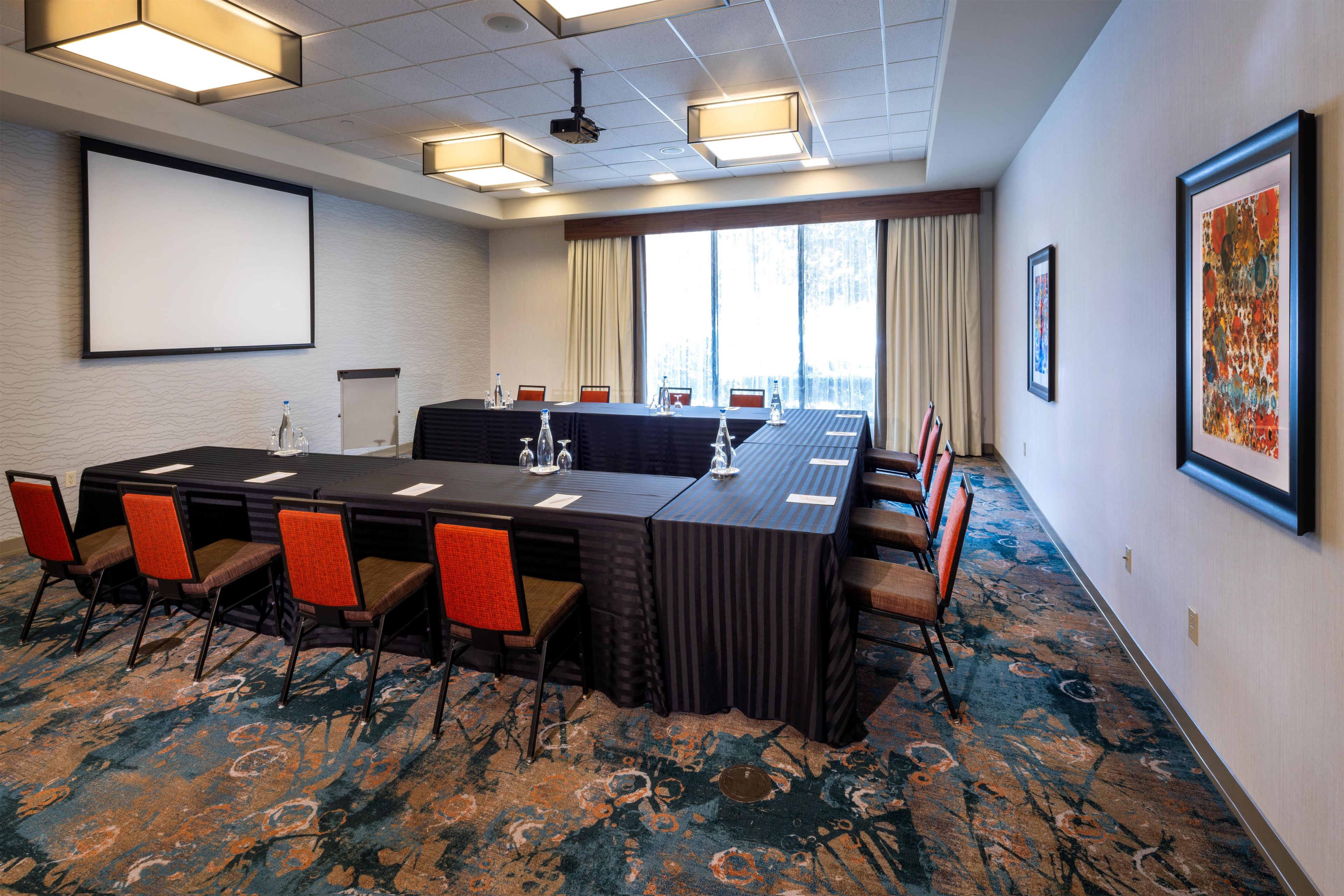 A conference room with a long table, chairs, a projector screen, and a large window with curtains.