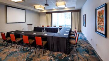 A conference room with a long table, chairs, a projector screen, and a large window with curtains.