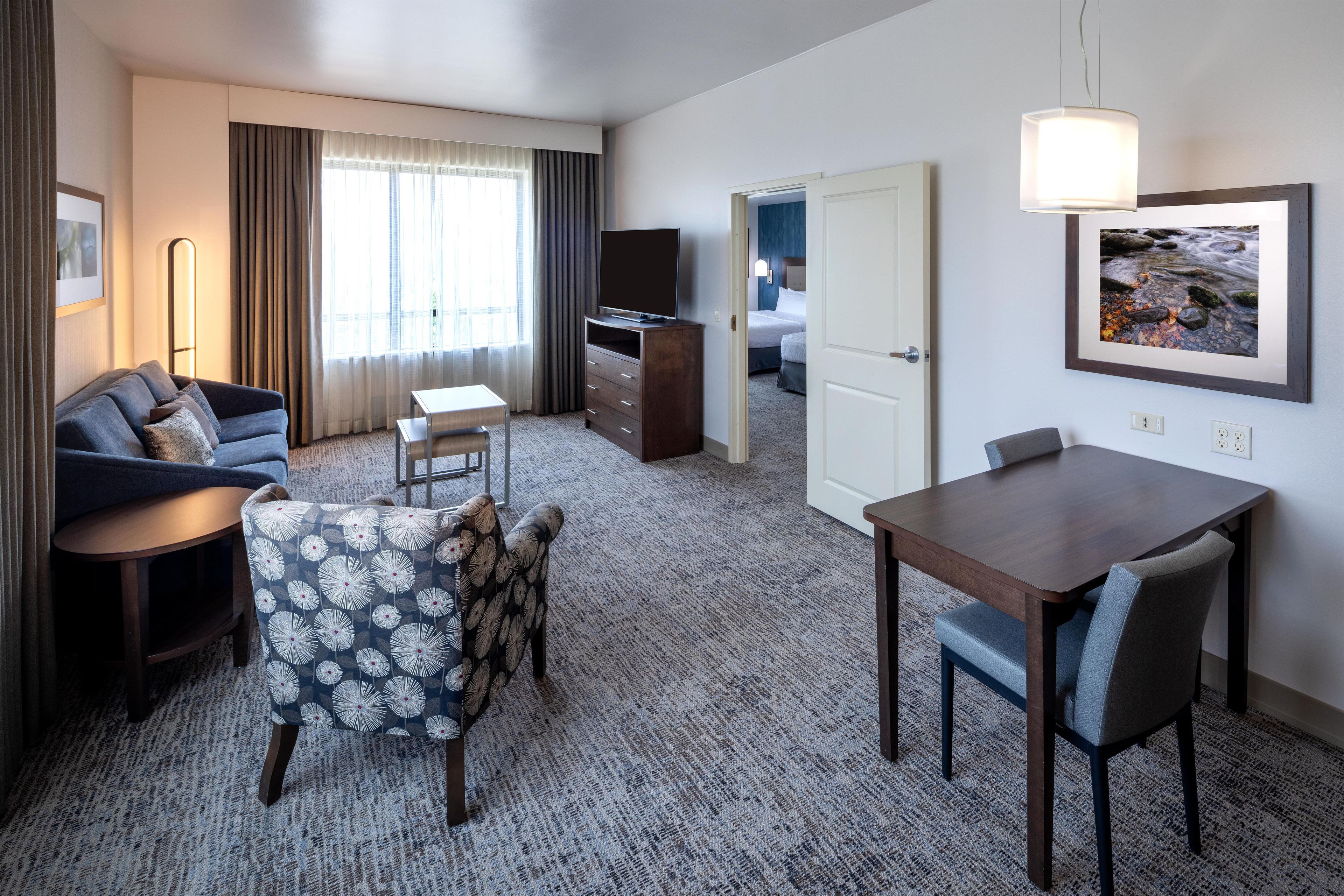 A hotel room with a sofa, a small table, a dining table with chairs, a TV, a bed, and a painting on the wall.