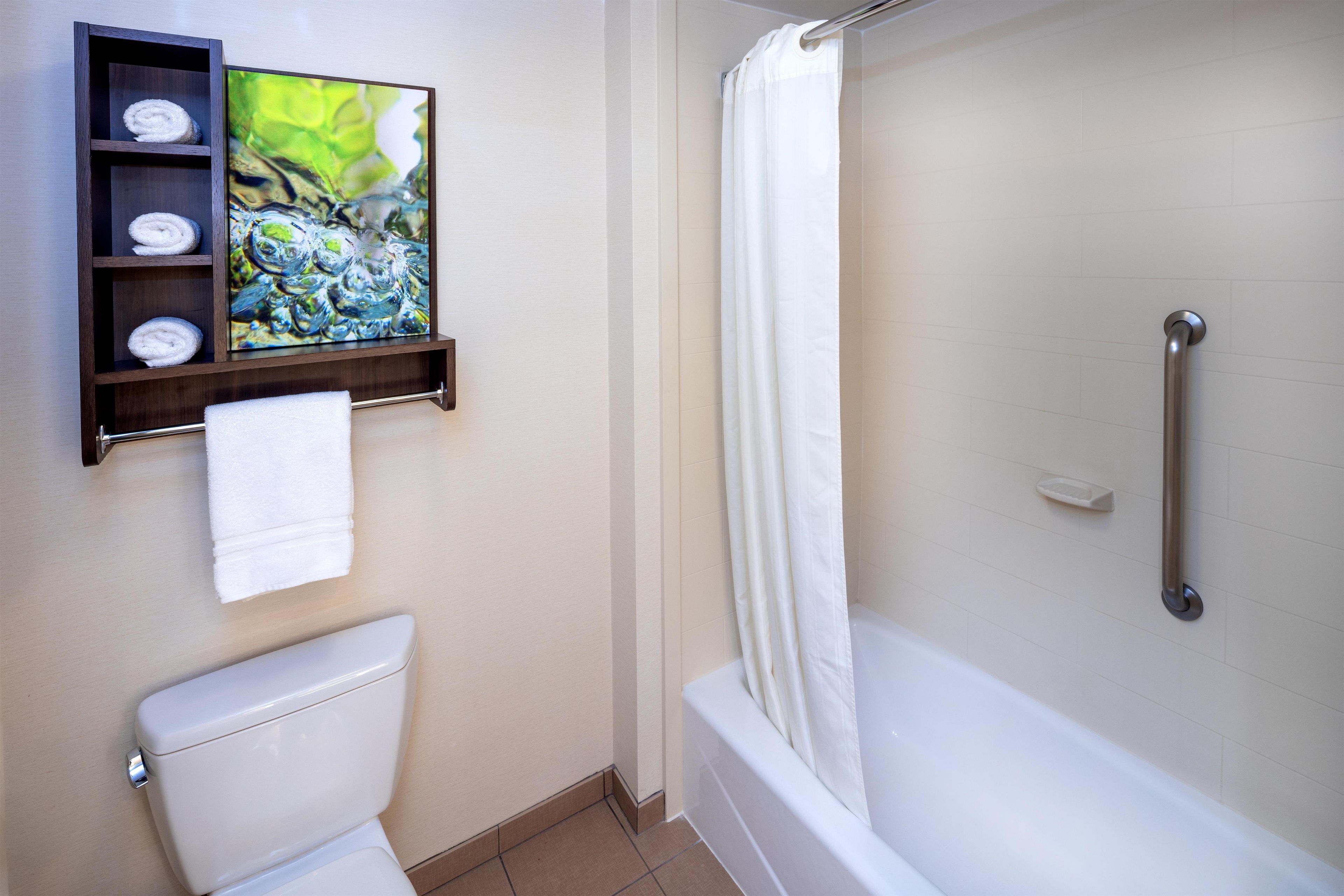A bathroom with a toilet, a shower with a grab bar, and a shelf with towels and a painting.