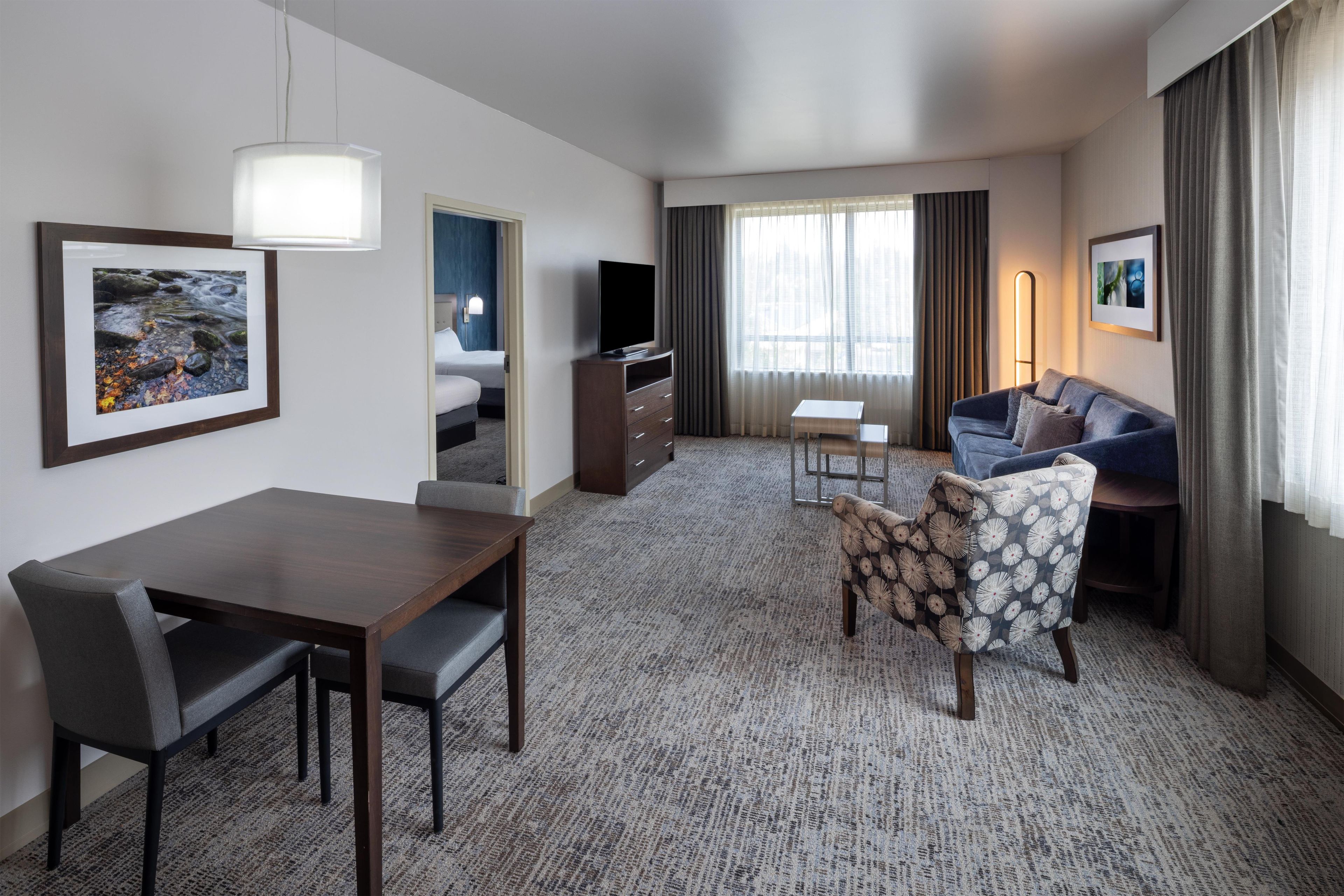 A hotel room with a dining table, a sofa, a TV, and a large window with curtains.