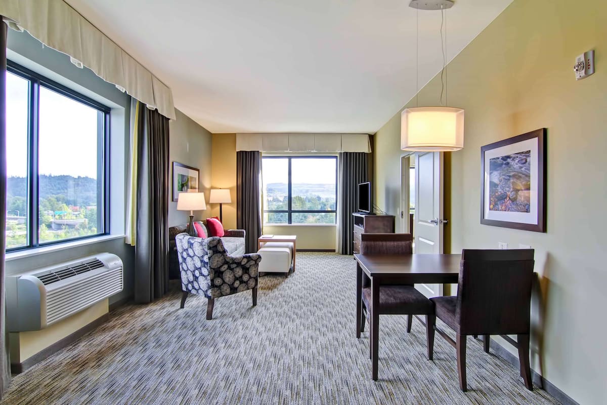 A hotel room with a large window, a sofa, a dining table, and a TV.