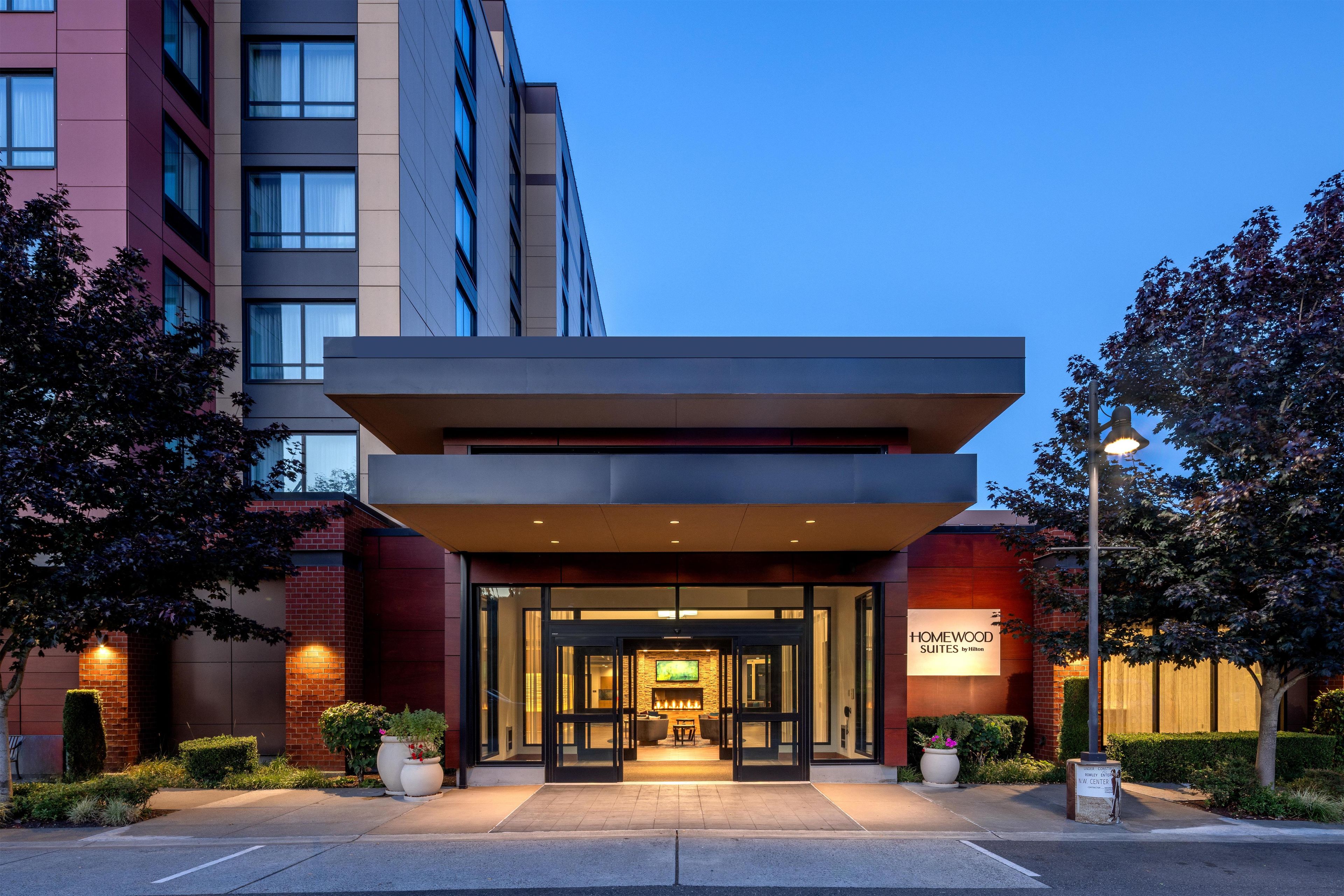 Entrada de hotel com design moderno, apresentando uma porta de vidro e uma placa com a inscrição 'Homewood Suites by Hilton'.