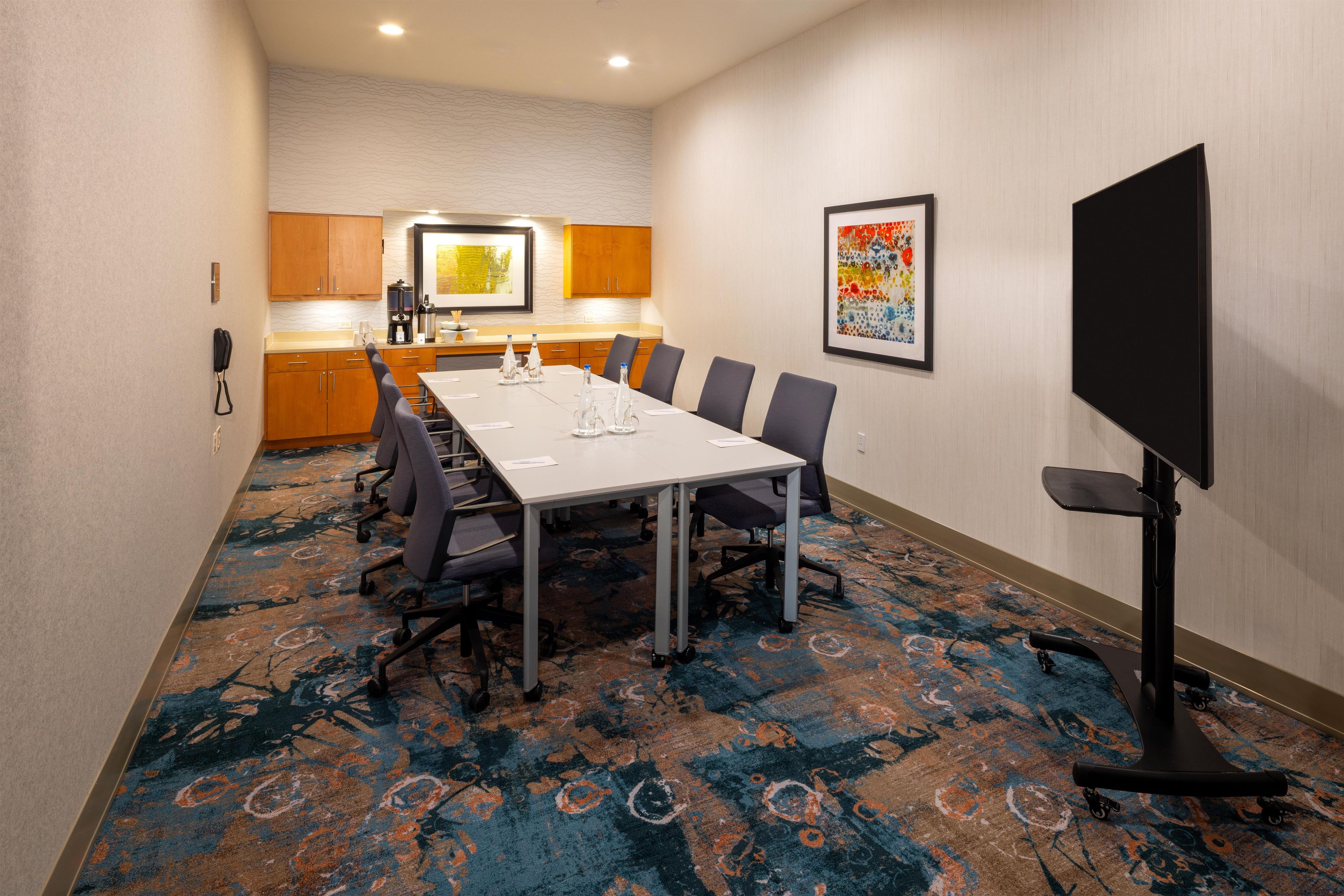 A conference room with a long table, chairs, a projector screen, and a kitchen area in the background.