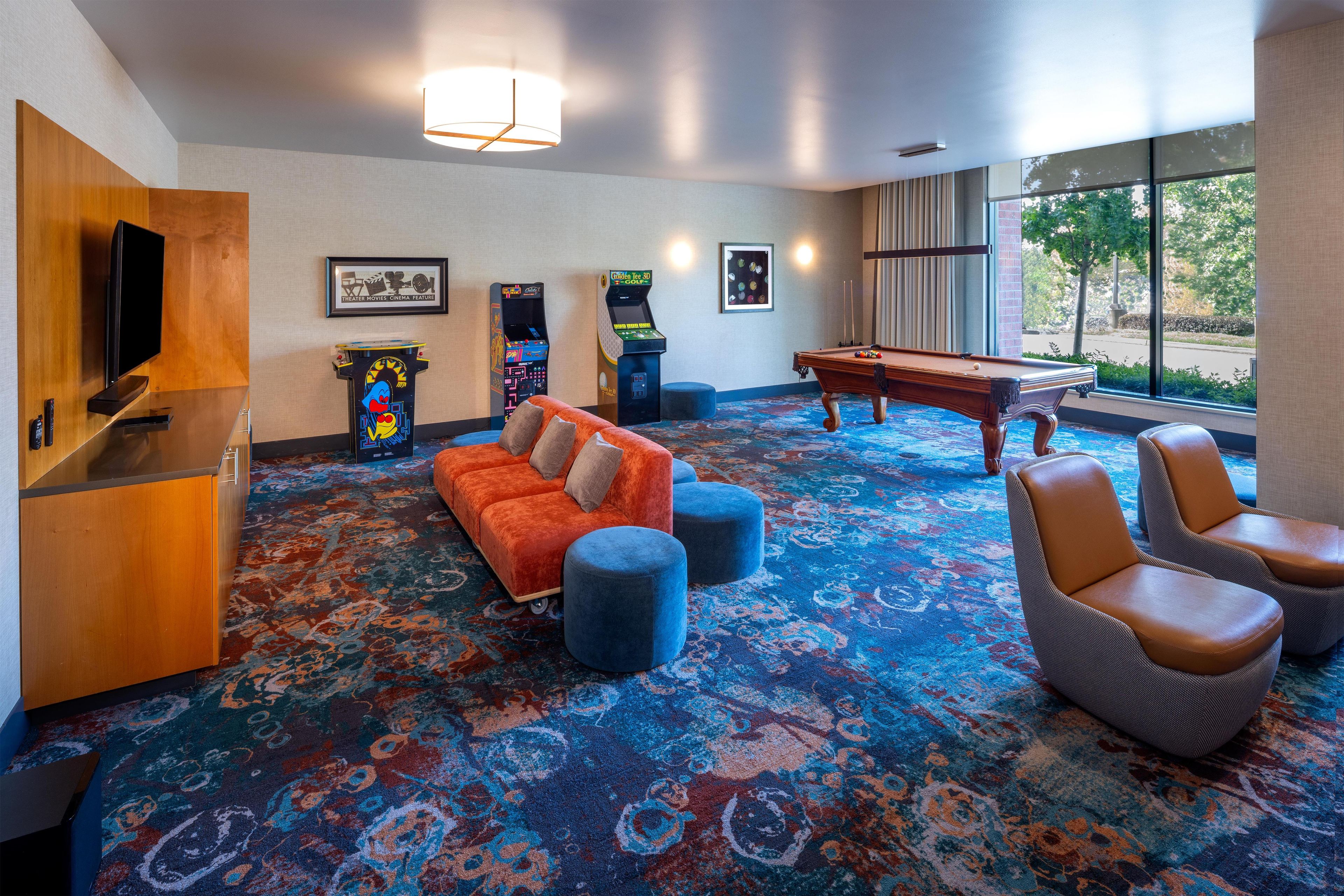 A hotel room with a pool table, a sofa, a television, arcade games, and a large window with a view.