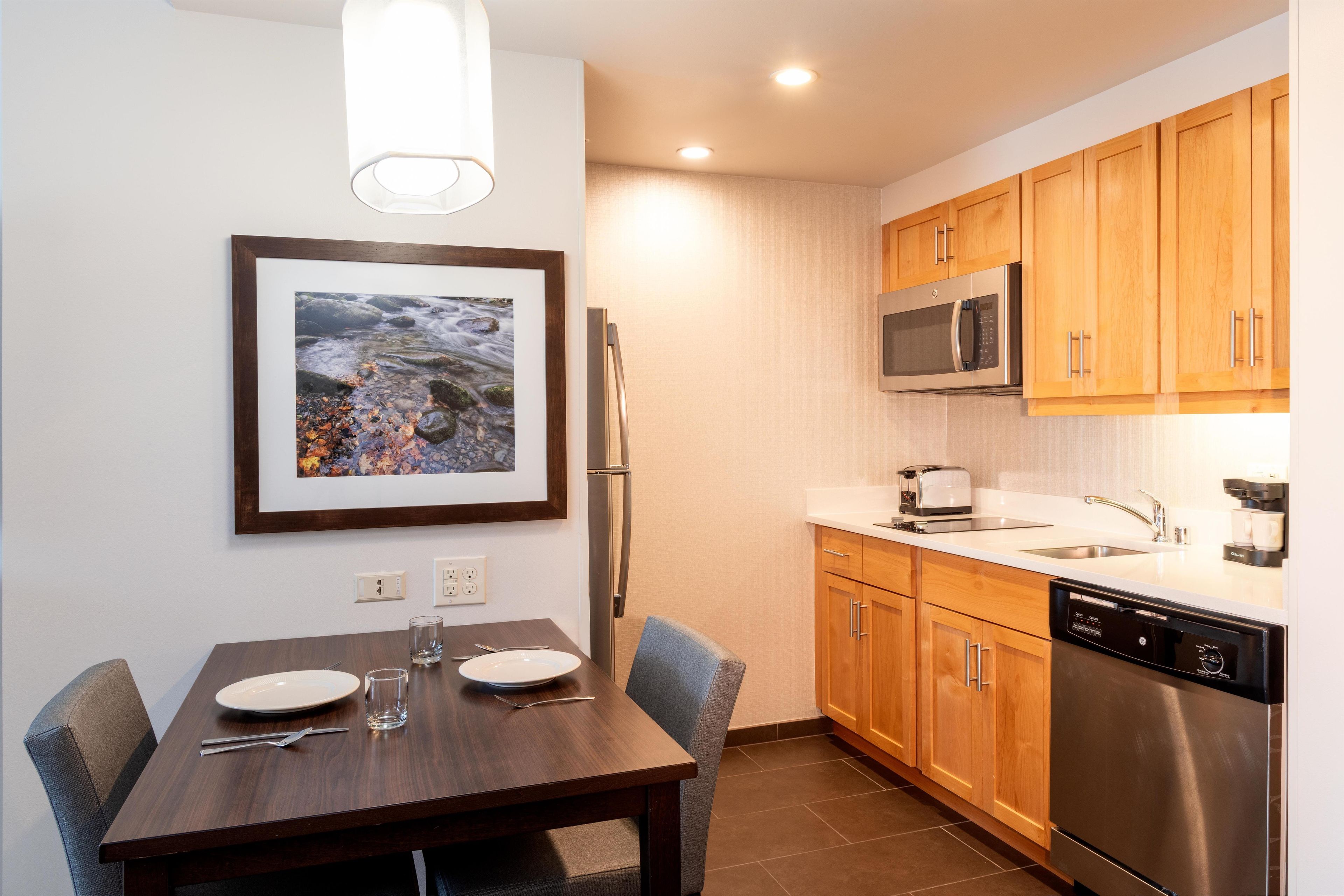 A modern kitchen with wooden cabinets, a stainless steel refrigerator, a microwave, and a dishwasher. There is a dining table with chairs and a framed picture on the wall.