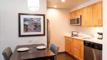 A modern kitchen with wooden cabinets, a stainless steel refrigerator, a microwave, and a dishwasher. There is a dining table with chairs and a framed picture on the wall.