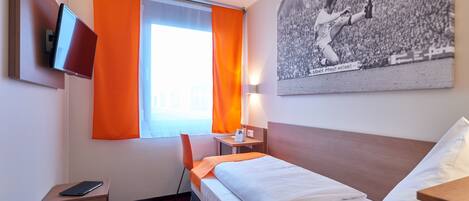 A hotel room with a bed, a desk with a chair, a TV mounted on the wall, and a window with orange curtains.
