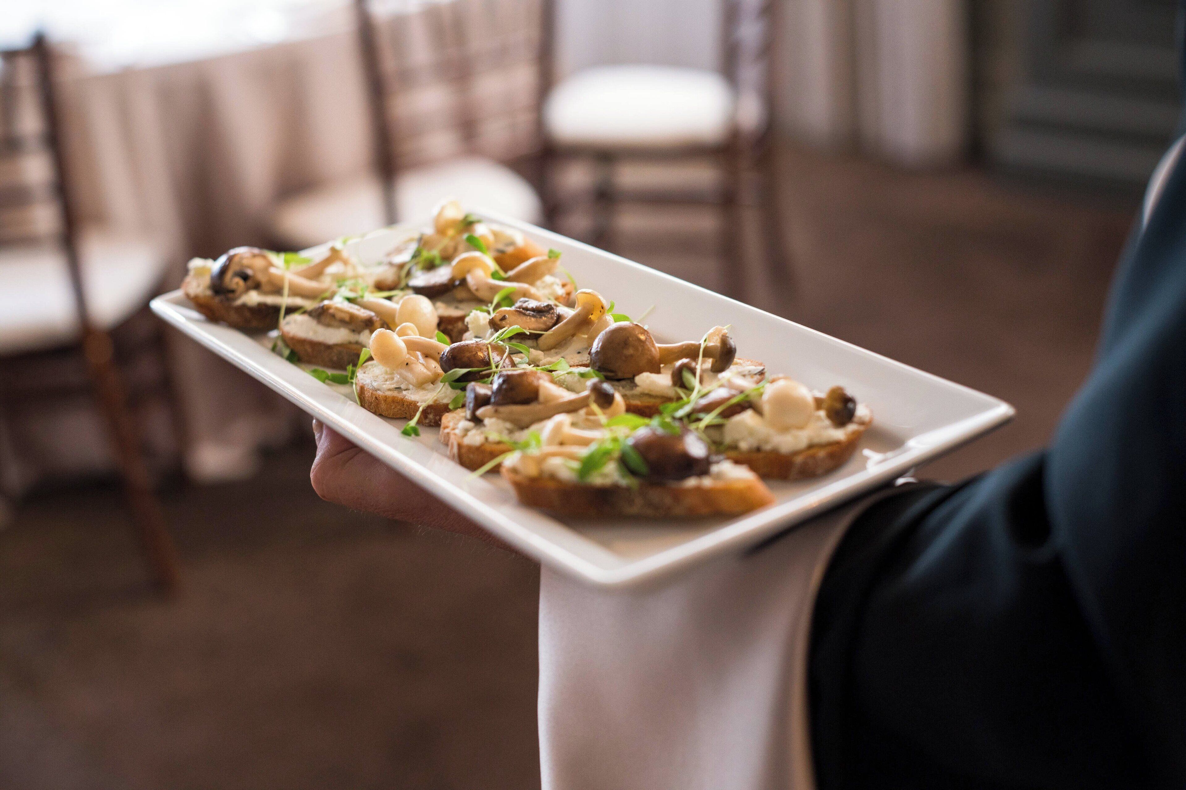 A person holding a rectangular white plate with food items, likely appetizers, in a setting with wooden chairs and a neutral-toned interior.