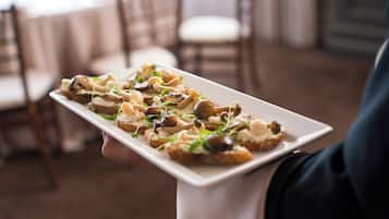 A person holding a rectangular white plate with food items, likely appetizers, in a setting with wooden chairs and a neutral-toned interior.