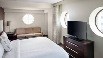 Una habitación de hotel con una cama grande, un sofá, un televisor sobre un aparador y dos ventanas redondas.