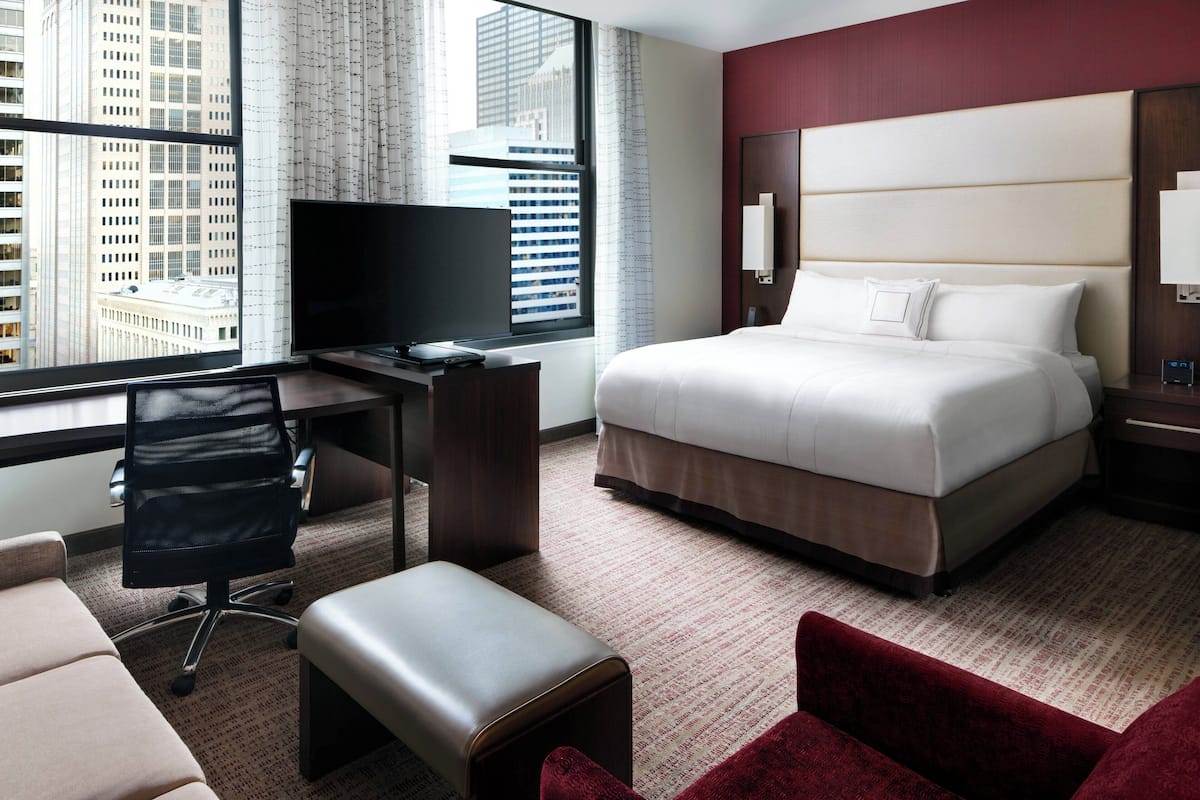 A hotel room with a large bed, a desk with a computer, a TV, and a view of city buildings.