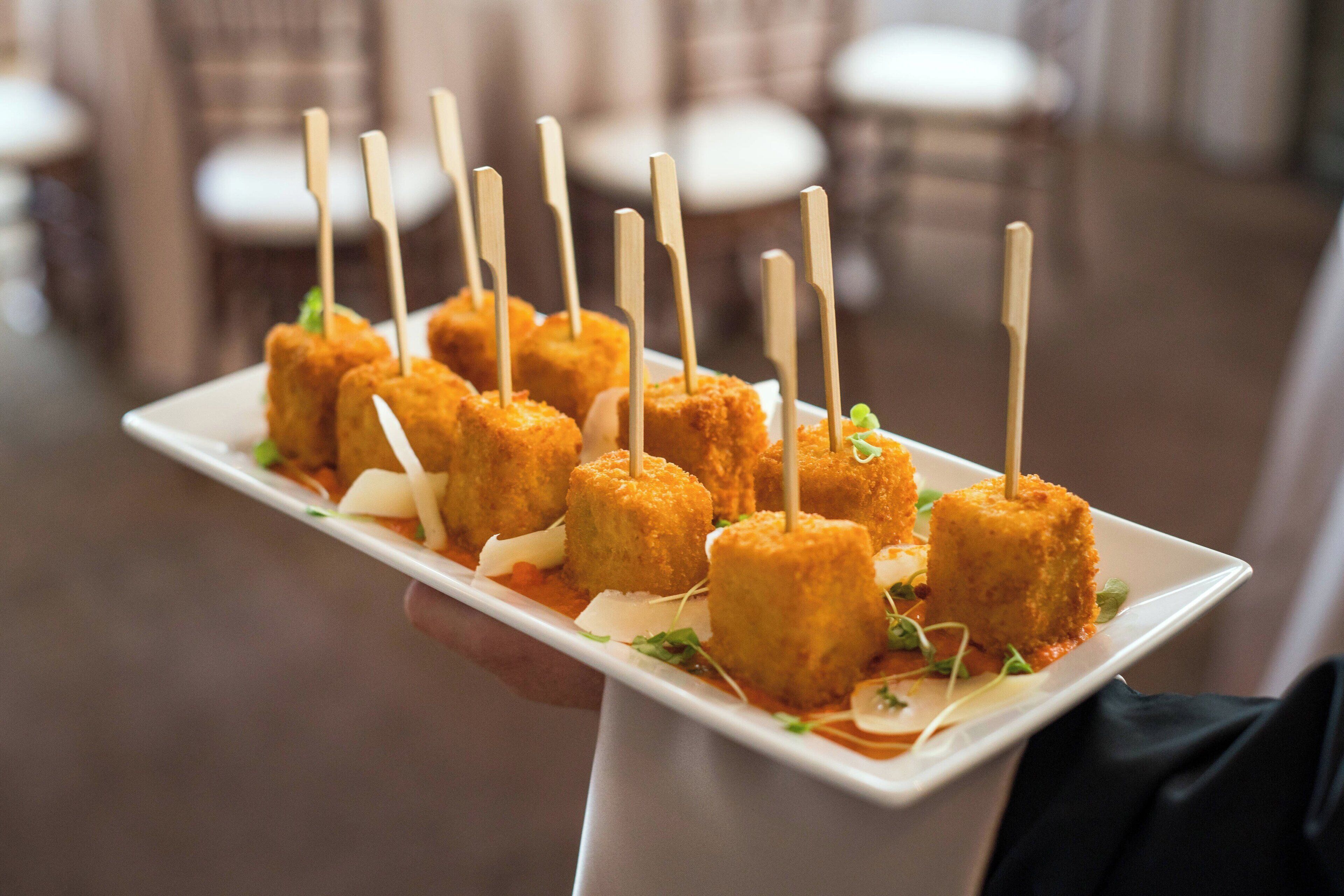 A white rectangular plate with several golden-brown, breaded items with toothpicks, garnished with green leaves and white shavings, held by a person in a black sleeve.