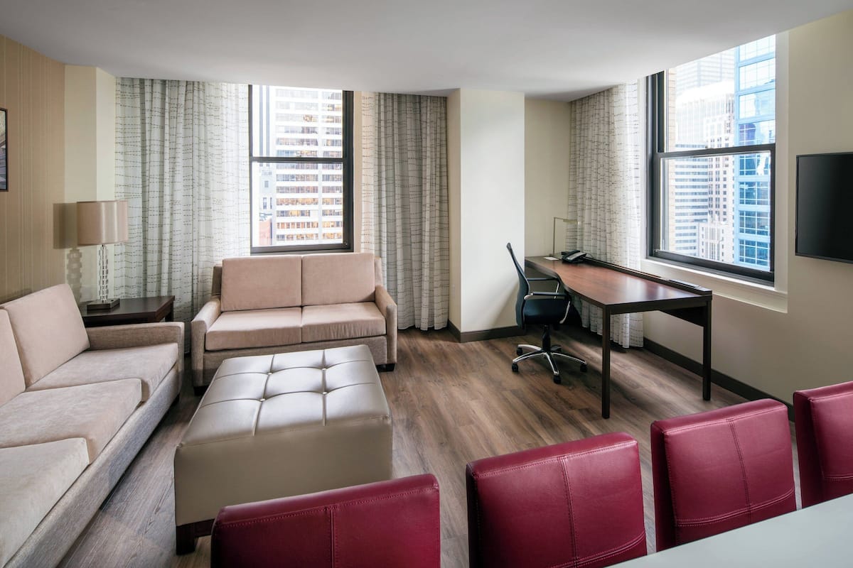 A modern hotel room with a sofa, ottoman, red chairs, a desk, and a TV.