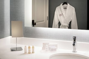 A modern bathroom with a white vanity, a mirror, a robe hanging on the wall, and various toiletries on the counter.