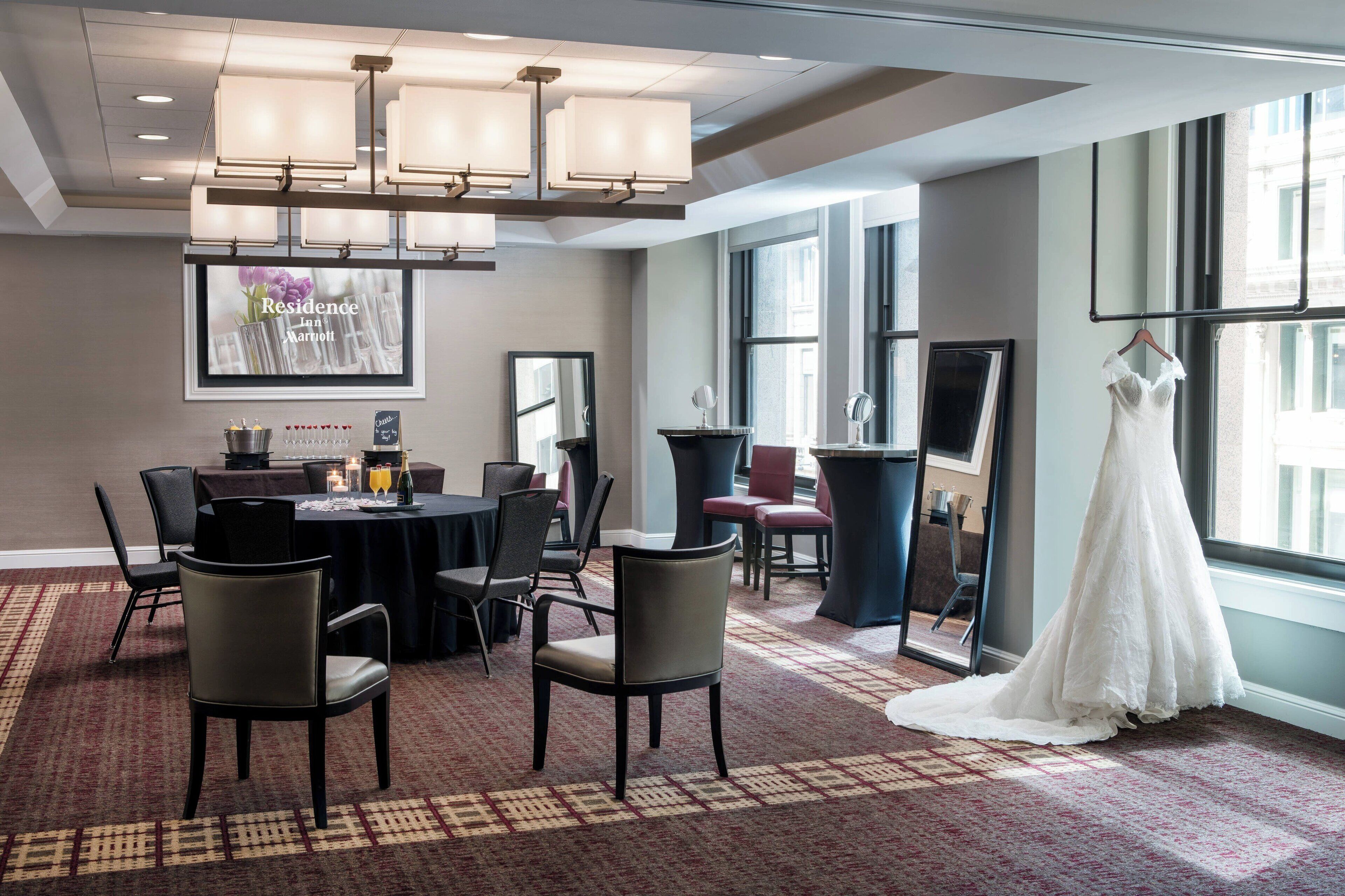 A hotel room with a large window, a chandelier, a round table with chairs, and a wedding dress hanging by the window.