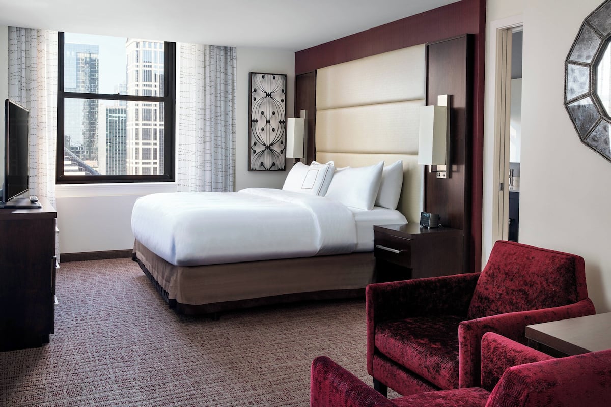 A hotel room with a large bed, a desk, a chair, a TV, and a window with a city view.