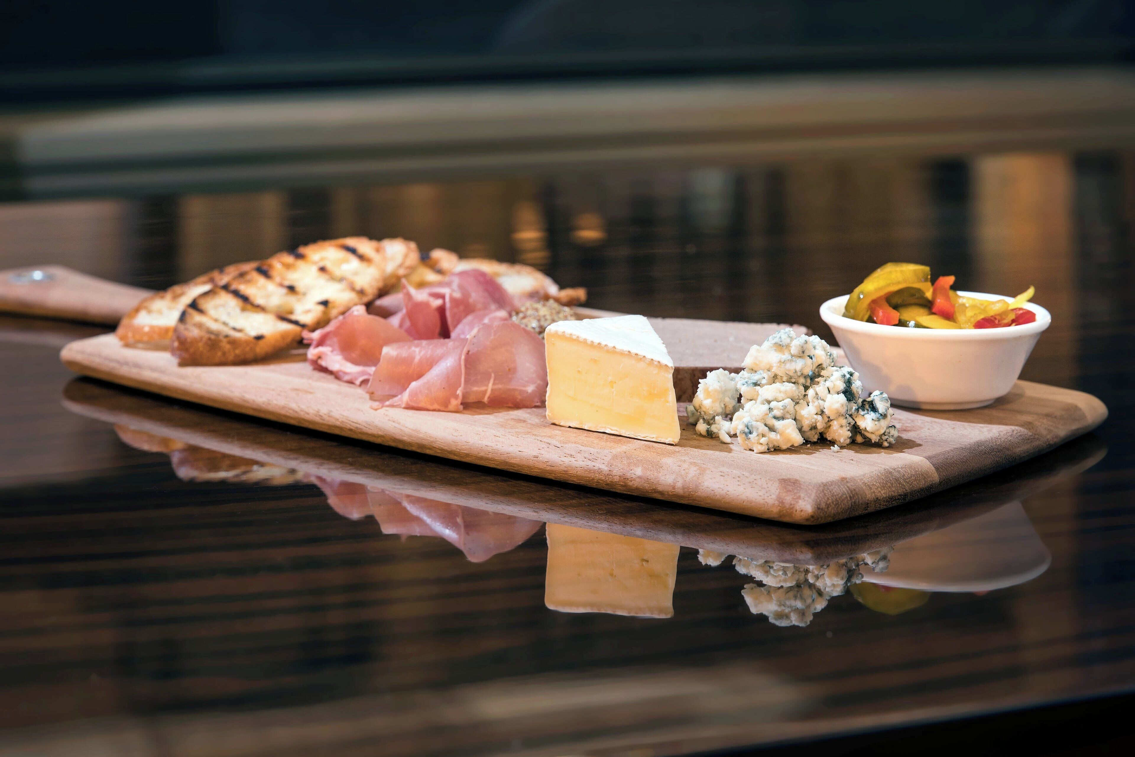 A wooden board with various cheeses, meats, and pickles on a reflective surface.