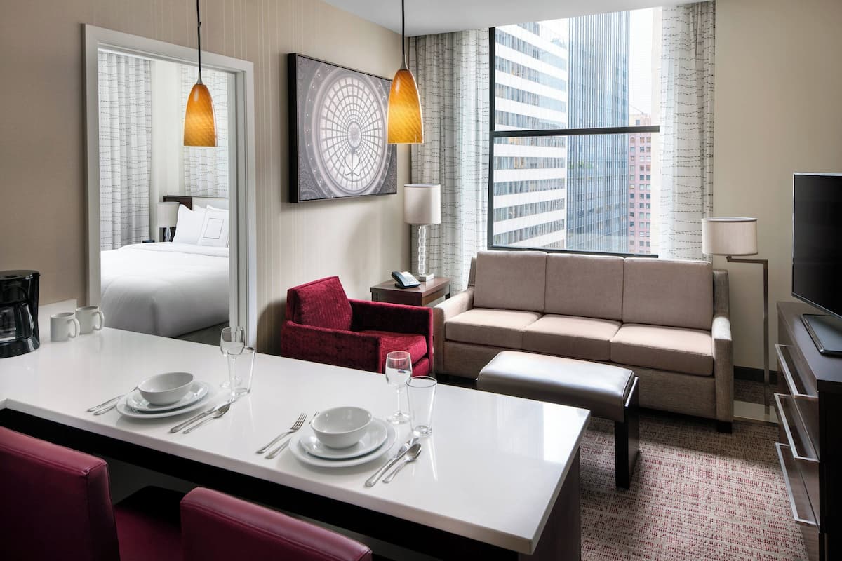 A hotel room with a dining area, a sofa, a TV, and a large window with a city view.