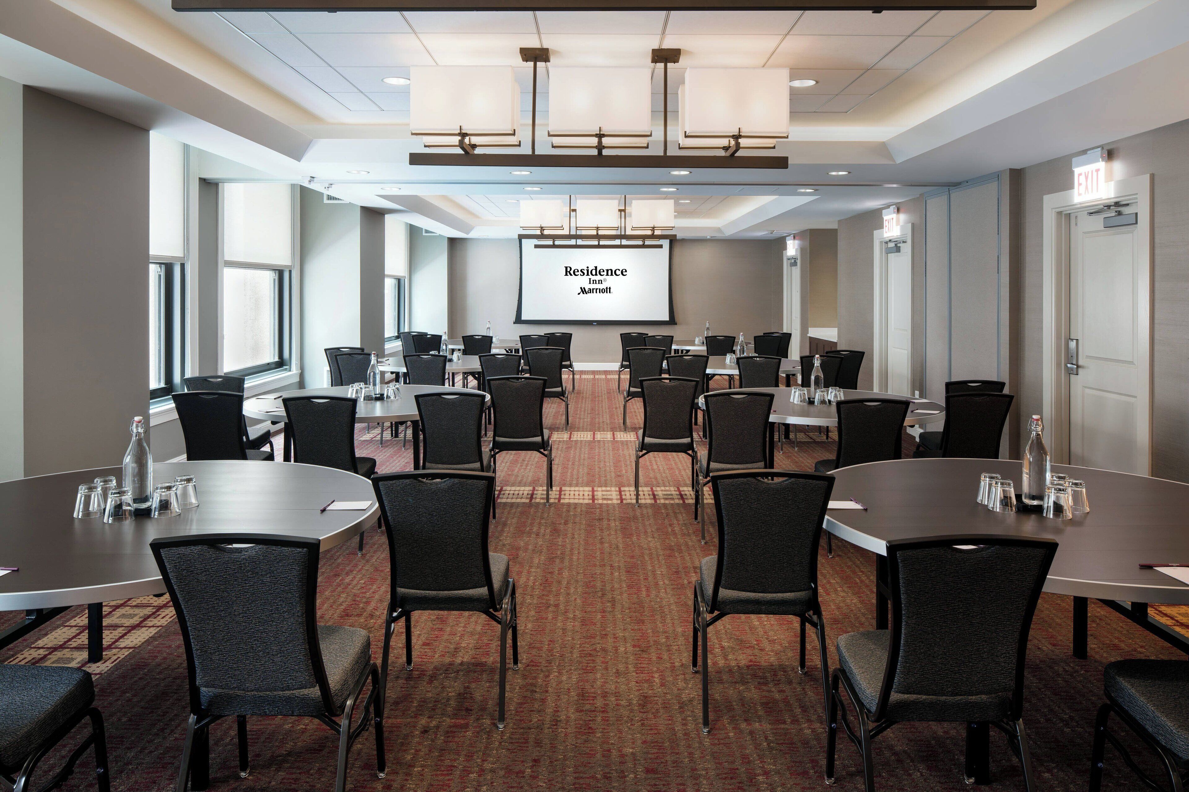 Un salón de conferencias con varias mesas y sillas, una pantalla grande mostrando 'Residence Inn Marriott' y señales de salida en las paredes.