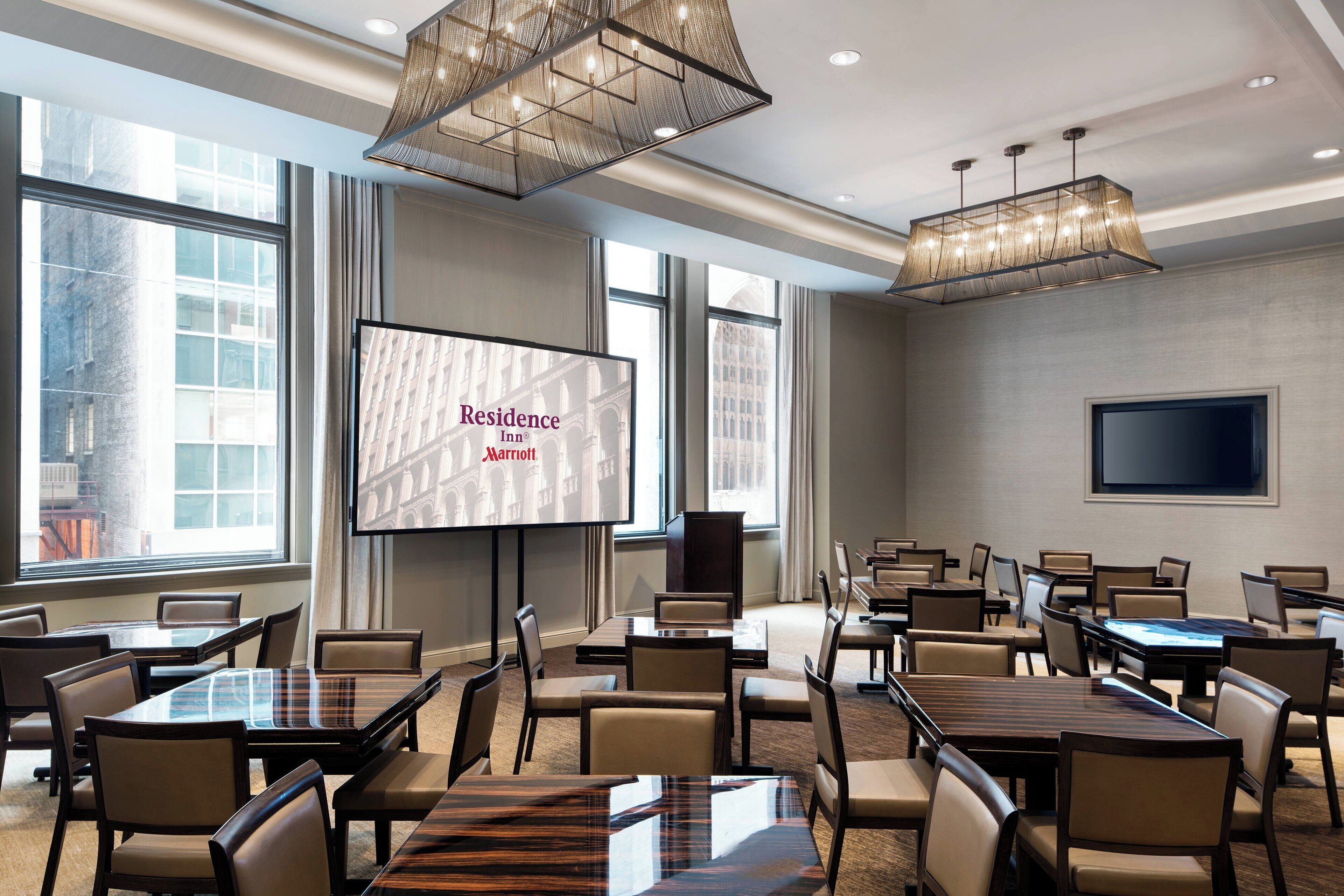 Un salón de conferencias con varias mesas y sillas, una pantalla grande mostrando 'Residence Inn Marriott' y amplias ventanas con vistas a la ciudad.