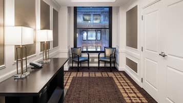 A hotel lobby with a reception desk, two chairs, a patterned carpet, and a view of a building through a large window.