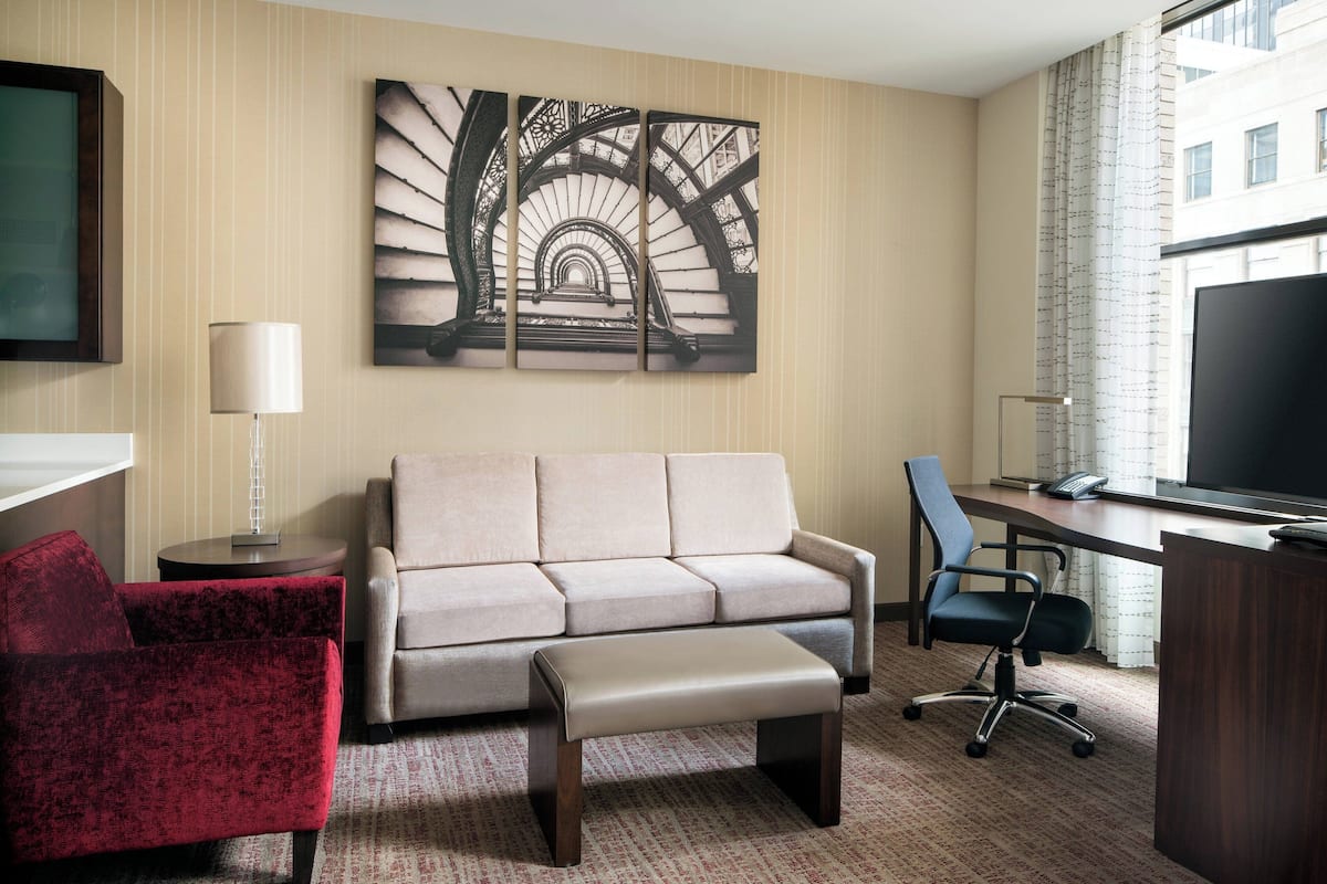 A hotel room with a sofa, a red armchair, a desk with a chair, a flat-screen TV, and a large abstract painting on the wall.