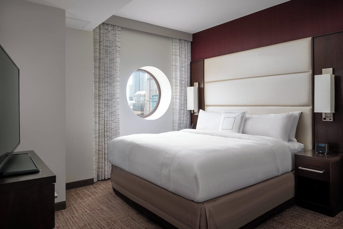 A hotel room with a large bed, a round window, a nightstand, and a wall-mounted TV.