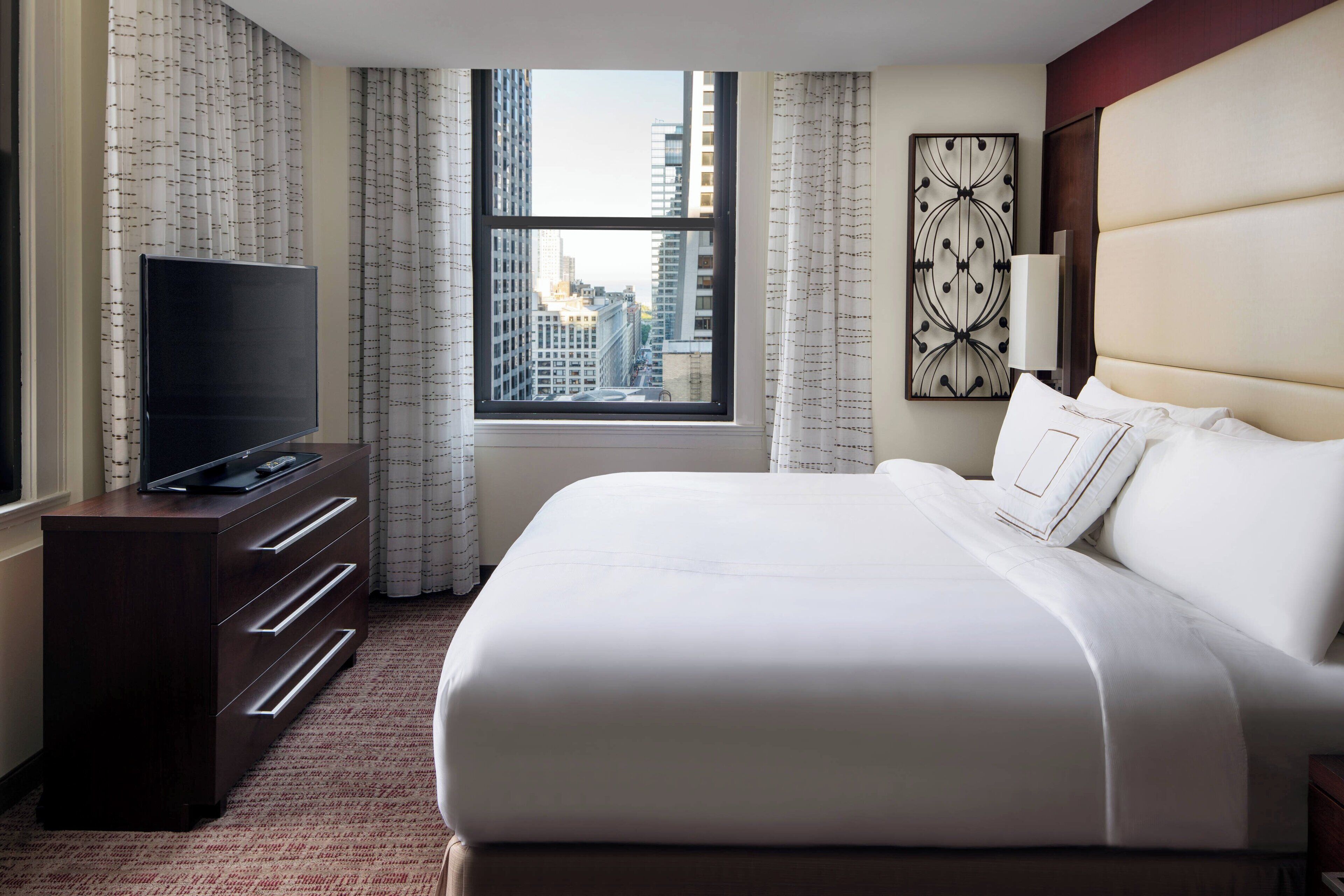 A hotel room with a large bed, a flat-screen TV on a wooden dresser, a window with a city view, and a decorative wall piece.