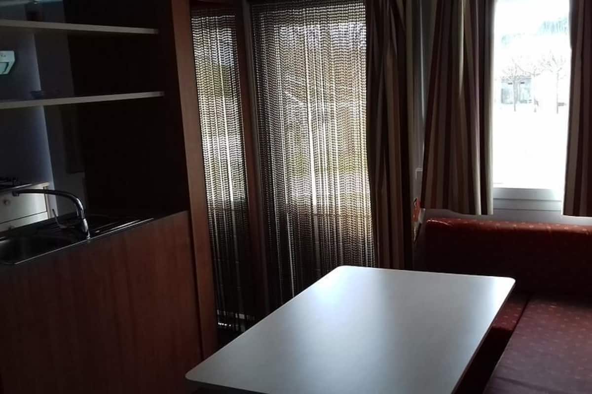 Una habitación compacta con kitchenette, una mesa de comedor, un banco y una ventana con cortinas.