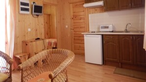 A wooden-paneled room with a wicker chair, a small table, a white refrigerator, a microwave, and wooden cabinets. - Camping Lluçanés (Olost)