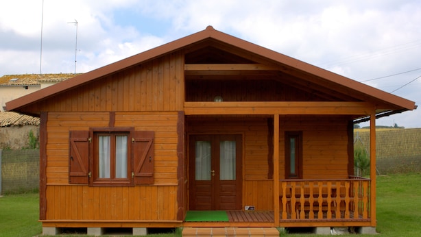 A two-story wooden house with a balcony, a front door, and a green mat at the entrance. - Camping Lluçanés (Olost)