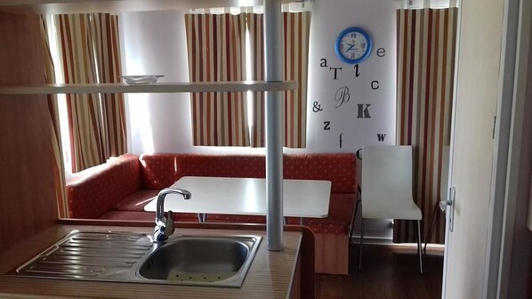 A compact kitchenette with a sink, a small table, and a chair. There is a wall clock and a partial view of a window with curtains.