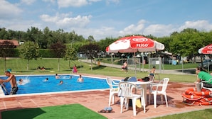 A swimming pool area with multiple people swimming and lounging, surrounded by white chairs and tables, and red umbrellas with the word "FRIGO" on them. - Camping Lluçanés (Olost)