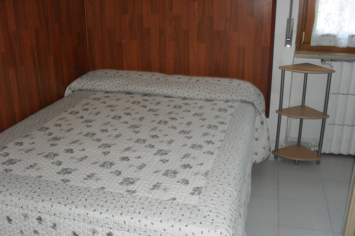A bedroom with a wooden wardrobe, a bed with a floral-patterned bedspread, and a metal shelf unit.