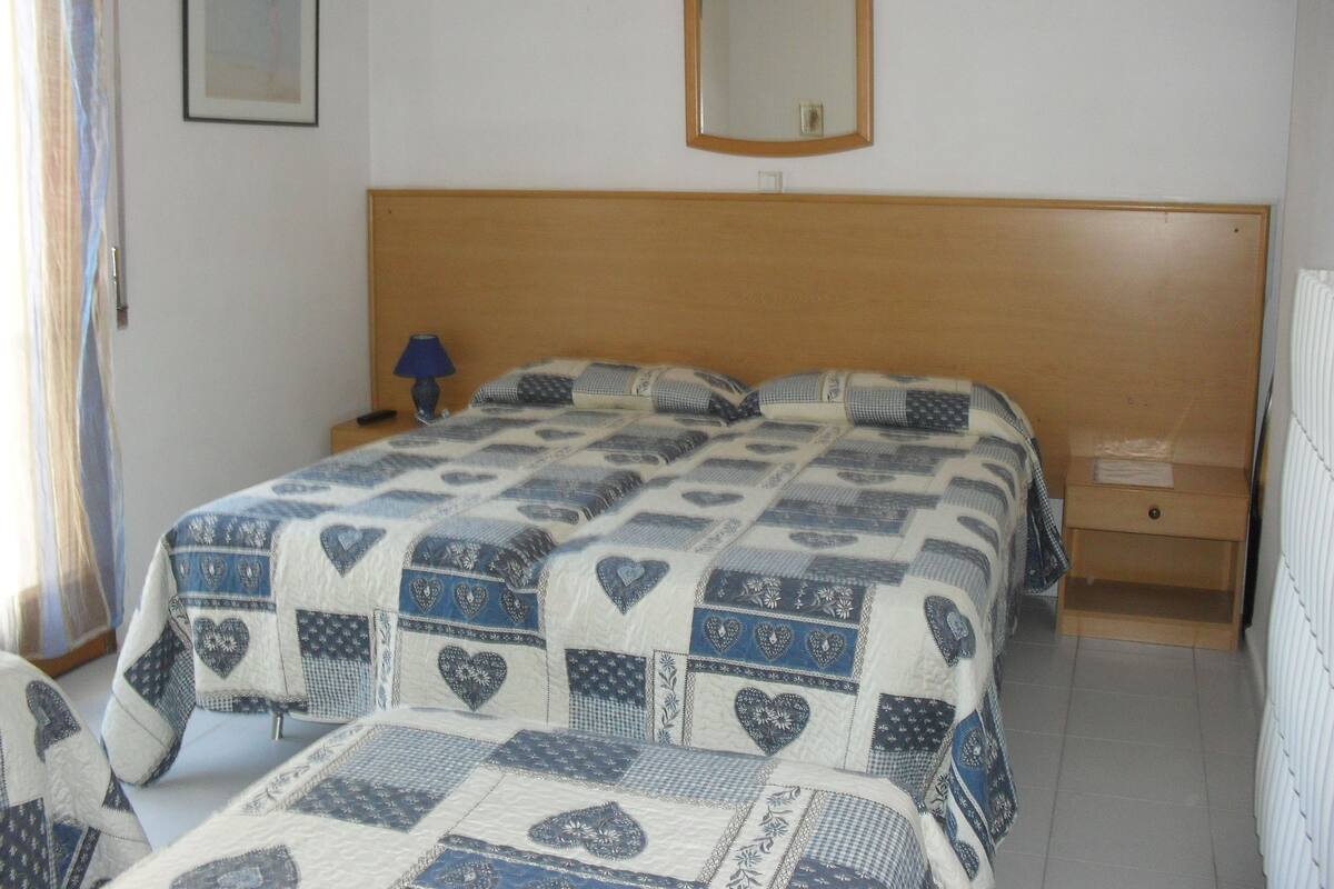 A bedroom with a wooden headboard, a bed with a patterned quilt, a bedside table with a lamp, a framed picture on the wall, and a mirror above the bed.