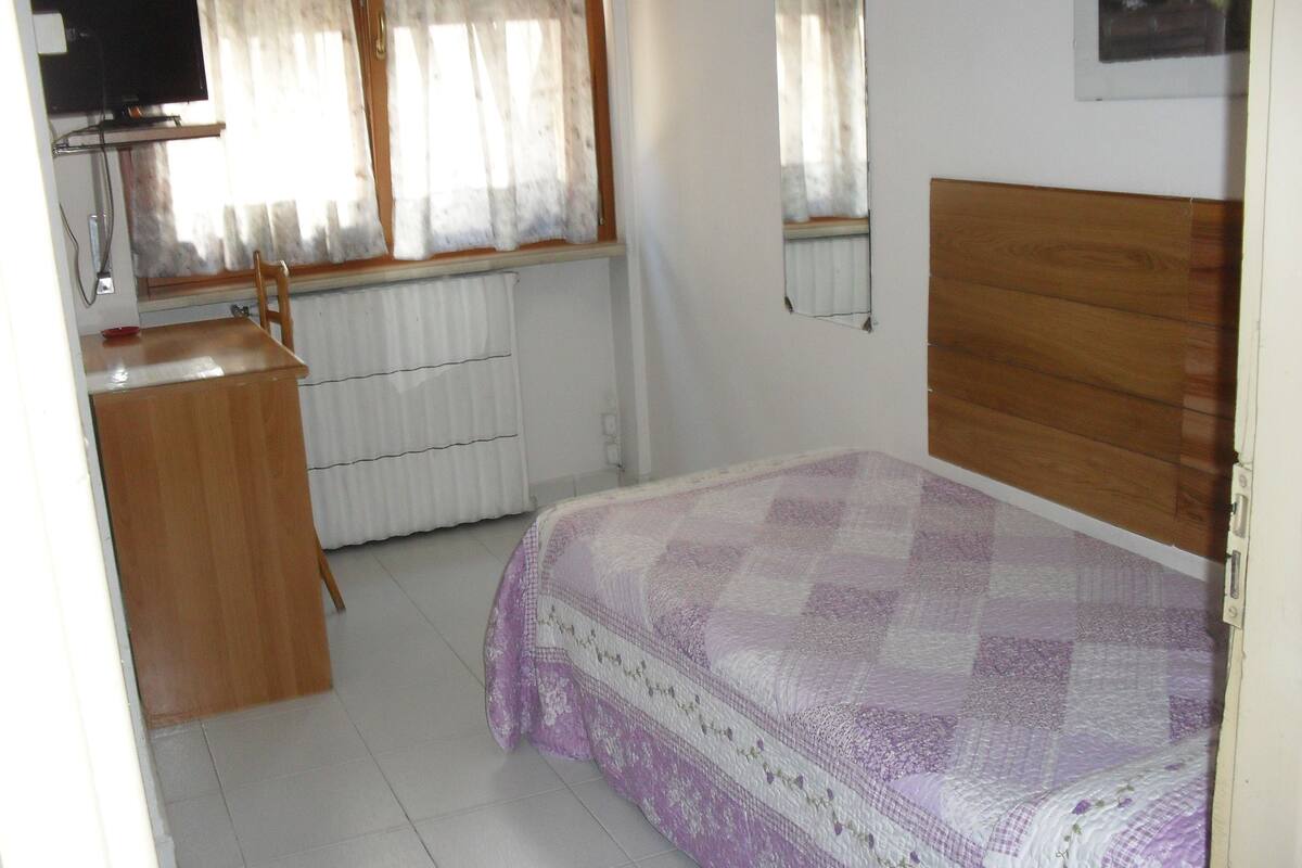 A bedroom with a bed, a wooden dresser, a radiator, and a television on a stand.