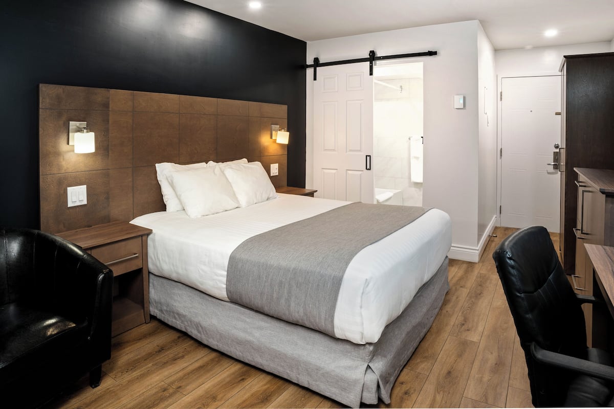 A modern hotel room with a large bed, wooden headboard, bedside lamps, a desk, and a chair.